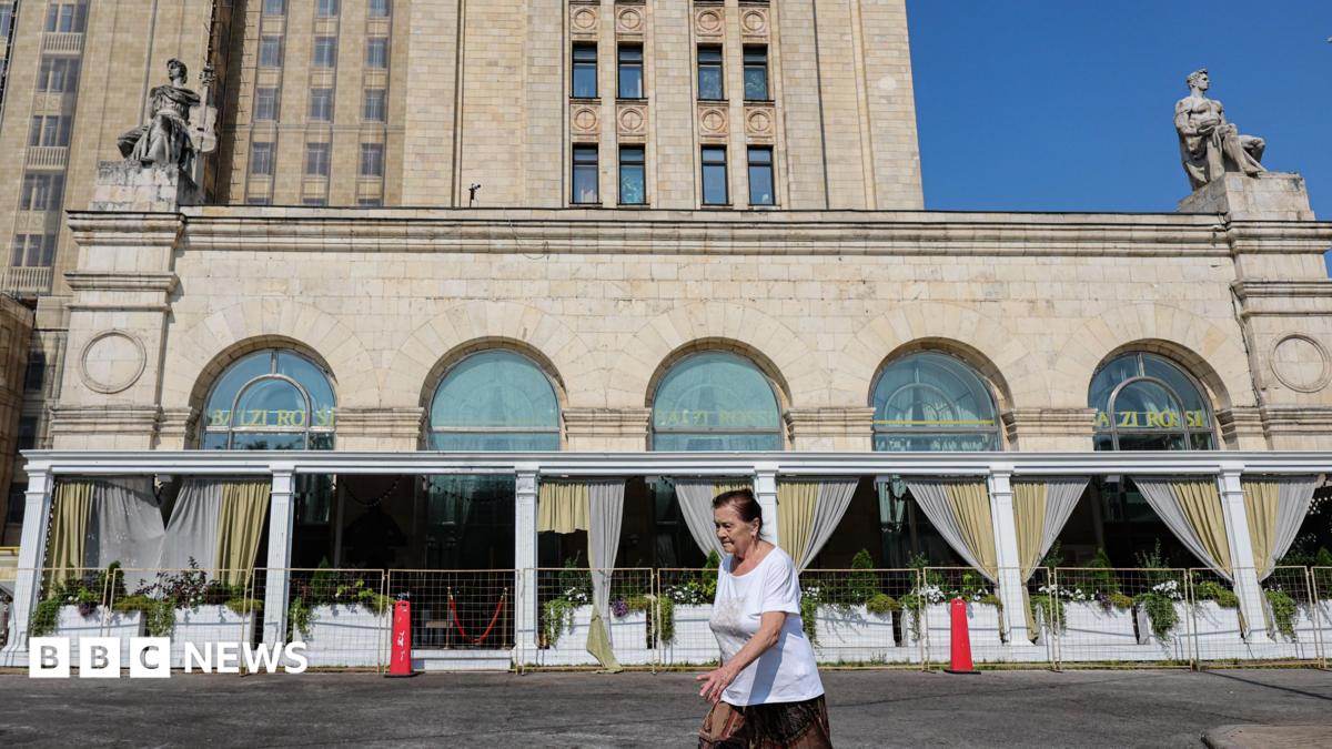 A woman wearing a red jumper and sunglasses, as well as a little bag on her right shoulder, talks on the phone in front of an exterior view of the Central Bank of the Russian Federation building on top of which the Russian flag can be seen
