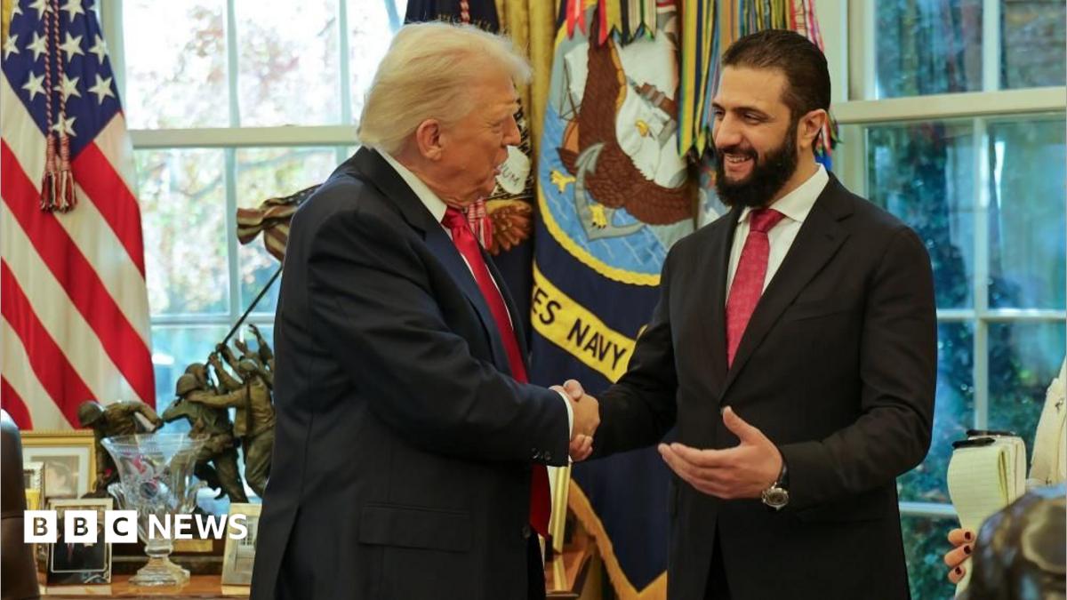 Two men in dark suits and red ties smile and shake hands in front of windows and American flags