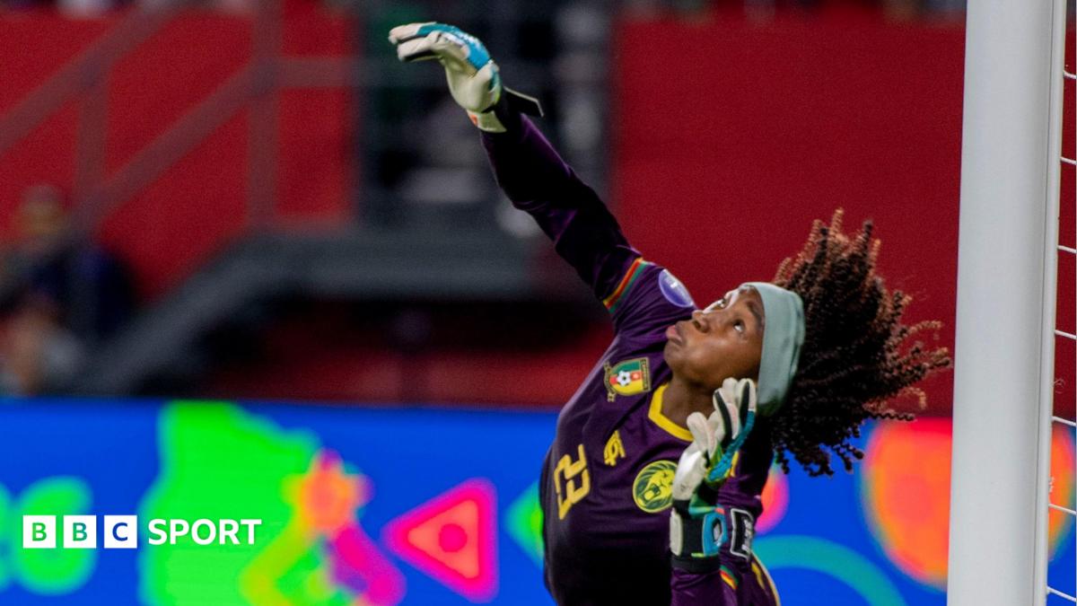 Cameroon goalkeeper Michaely Bihina, wearing an all-purple kit and wide blue headband, arches her back as she raises her right arm to push away a penalty