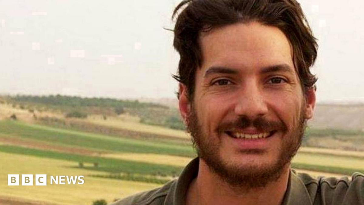 File image of Tice in front of a rural background. He has mid-length brown hair and a short brown beard and is wearing a khaki green polo shirt.
