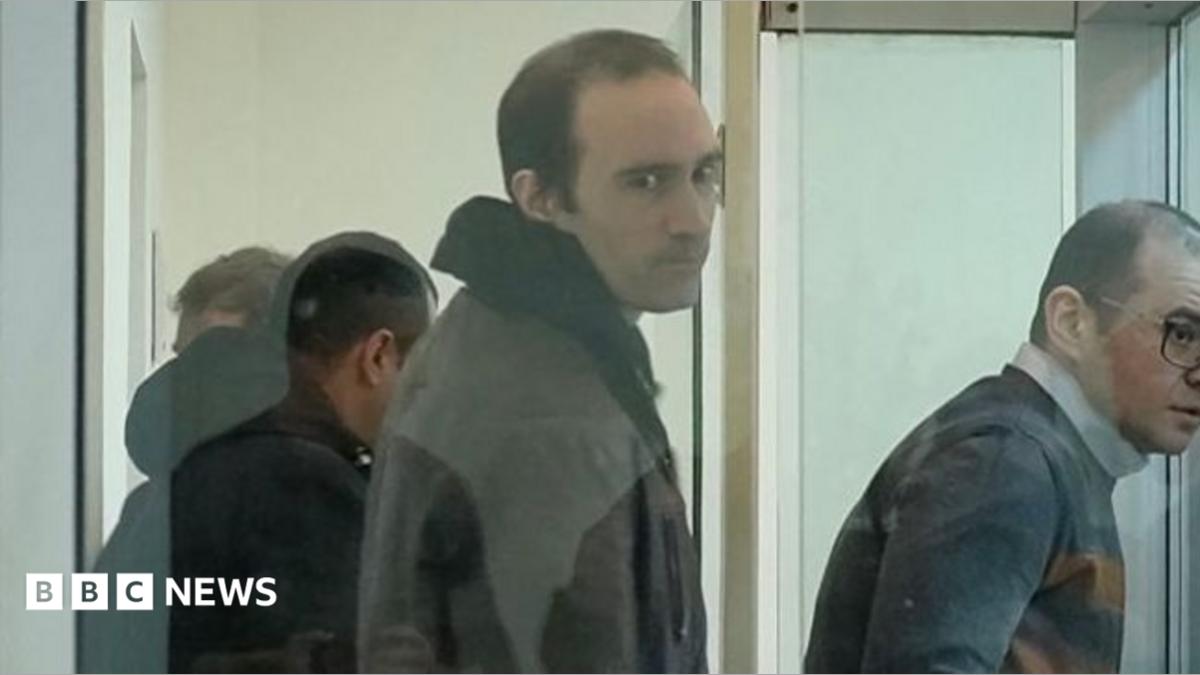 A screengrab from a video shows a man in a hoodie standing with short hair and a jacket and looking at the camera