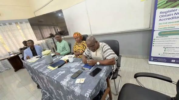 Five people seated at a long table in a conference room, with papers and mugs on the table, one man signing a document as others watch.
