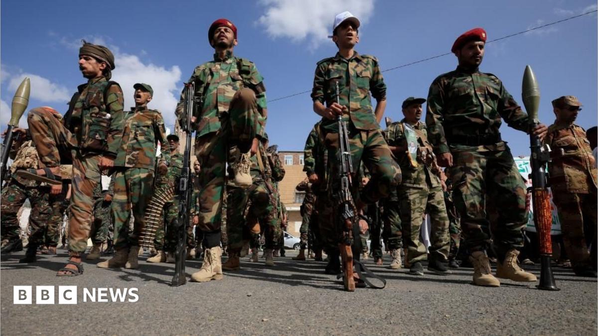 A dozen Houthi men wearing combat clothes, berets and holding large guns. The sky is blue and the men are looking straight ahead.