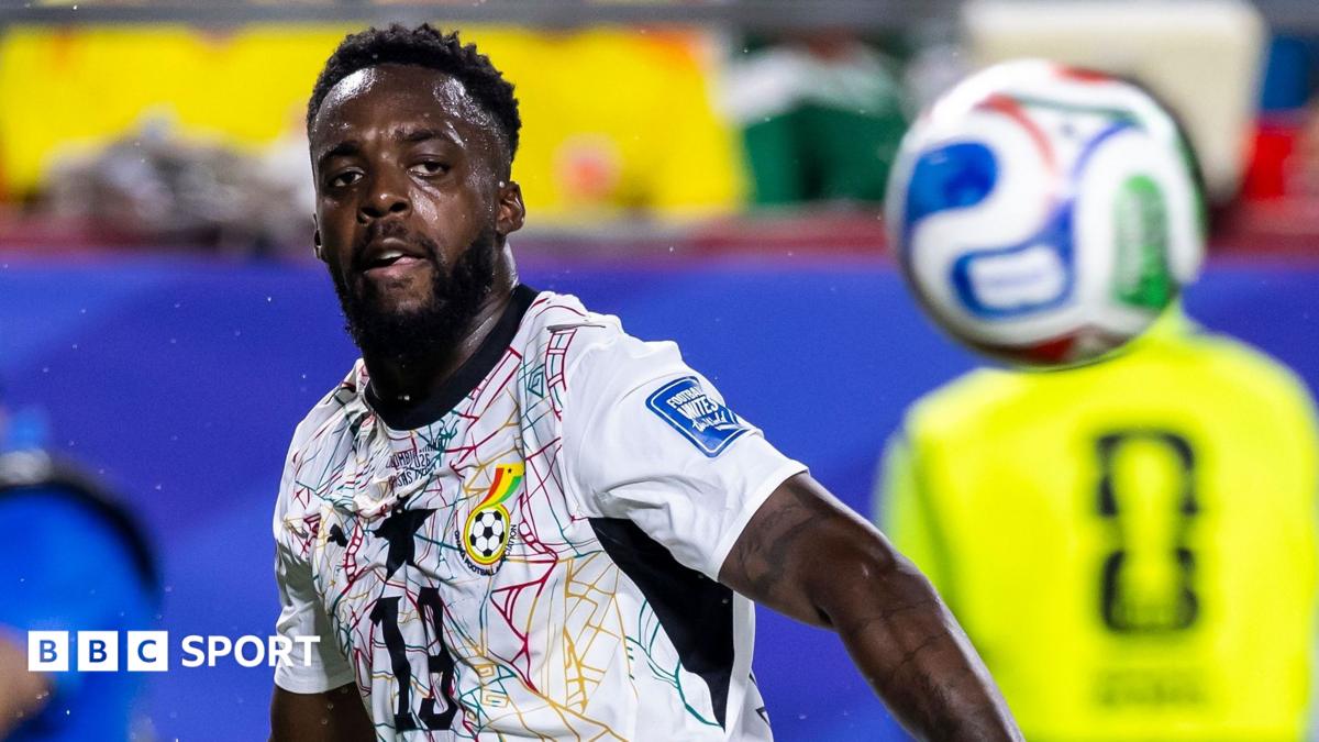 Inaki Williams, visible from stomach up wearing a white Ghana shirt with an intricate green, red and yellow spiderweb pattern on the chest, chases after a bouncing ball which is out of focus in the foreground during a match