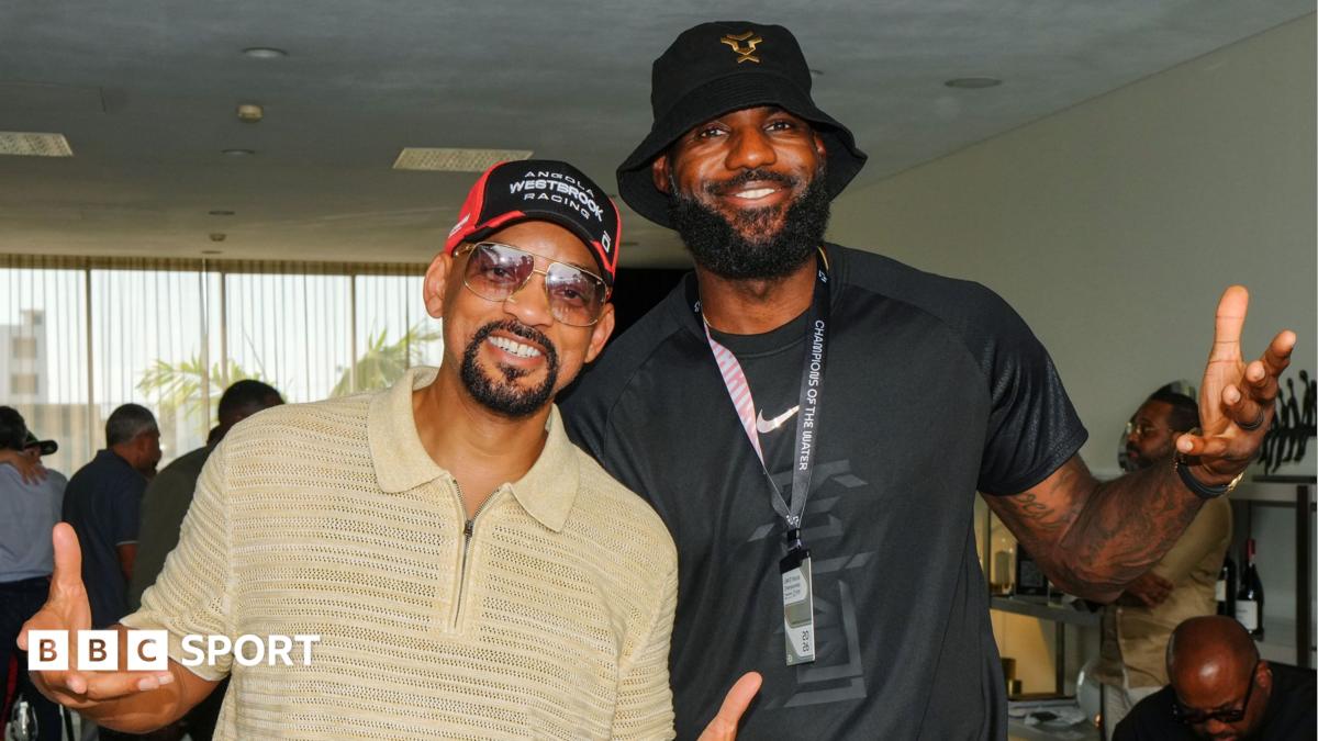 Will Smith, wearing a beige zip-up polo shirt, tinted sunglasses and a red and black baseball cap poses for a picture with LeBron James, who wears a black t-shirt and a black bucket hat. The two men, who are visible from waist up, are leaning in slightly towards each other, smiling and bending their arms at the elbow so that their forearms and hands are extended towards the camera in an expressive way with the palms of their hands open
