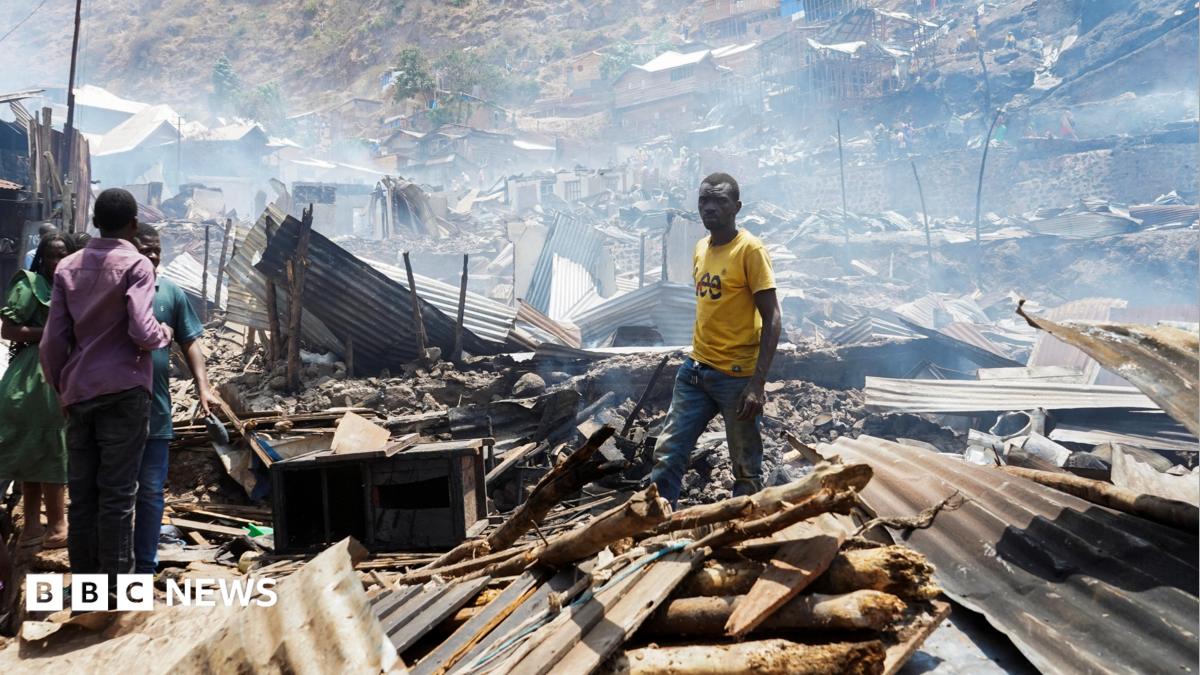 Man in yellow t-shirt stands in piles of rubble and corrugated iron while three other people stand on the left of the picture. The rubble seems to go on for some distance and the air is hazy.