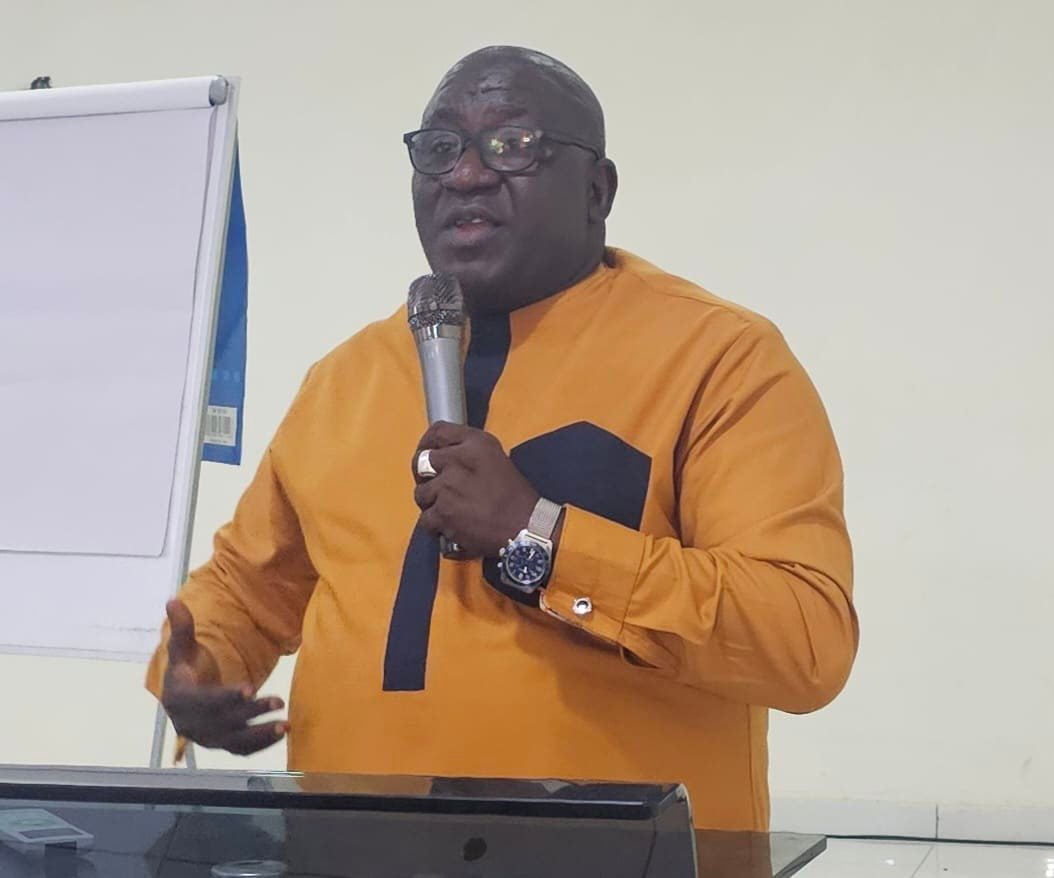 Man in an orange traditional shirt speaks into a microphone at a podium during a presentation.