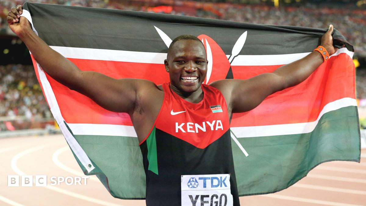 Faith Kipyegon, visible from chest up wearing an orange, yellow and green Kenyan zip-up running top, smiles and holds her left arm in front of her and stretches out four fingers in celebration. The vest has the word 'Kenya' written in white capital text across the chest while a Kenyan flag is on its left breast