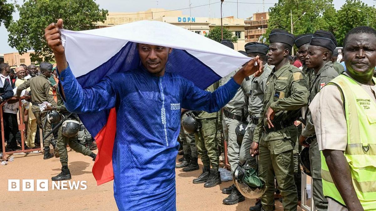 A man in a blue boubou drapes a Russian flag over his head. A group of people in army uniforms can be seen to the right of the picture.