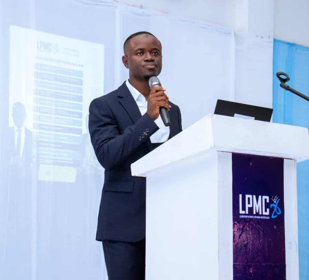 Man in a dark suit speaking into a handheld microphone at a white podium with a laptop, LPMC logo on the front panel.