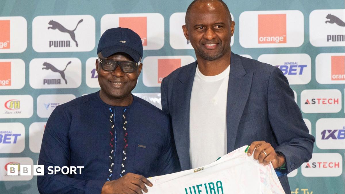 Senegalese Football Federation president Abdoulaye Fall and Patrick Vieria pose for photos at a press conference as between them they hold a white Senegal shirt with the name 'Vieira' in green on the back. Fall wears a dark blue baseball cap and matching dark blue robe while Vieira wears a grey-blue blazer over a cream t-shirt. Both men stand in front of a green hoarding bearing advertising logos