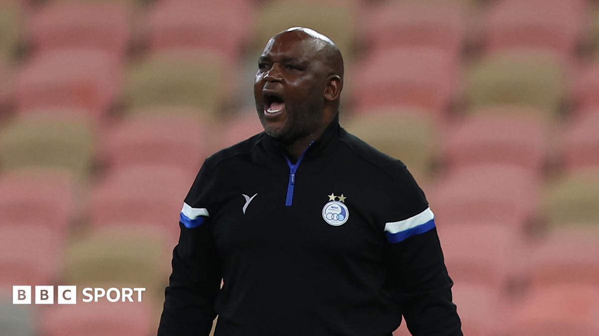 Pitso Mosimane, visible from waist up wearing a black Esteghlal tracksuit top with white and blue detailing on the sleeves, shouts during a match. In the background out of focus there is a stand of empty pink and tan seats