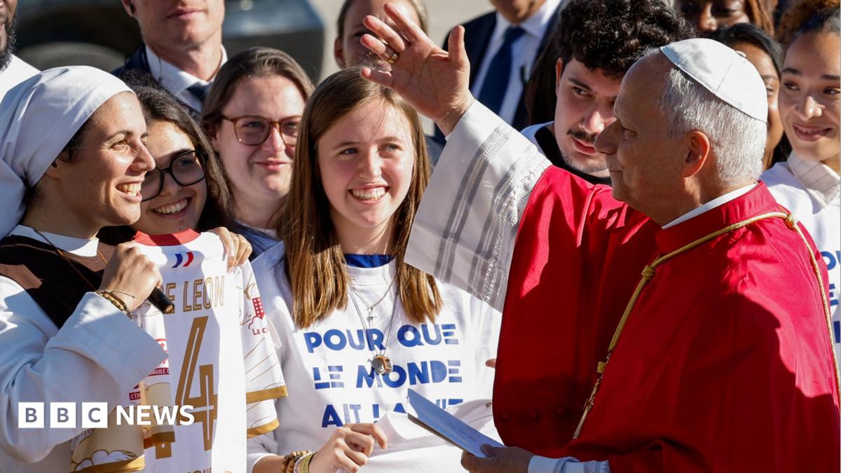 Pope in centre, with a waving Macron on his right and Brigitte Macron on his left. There is a French guard standing on either end of the picture.