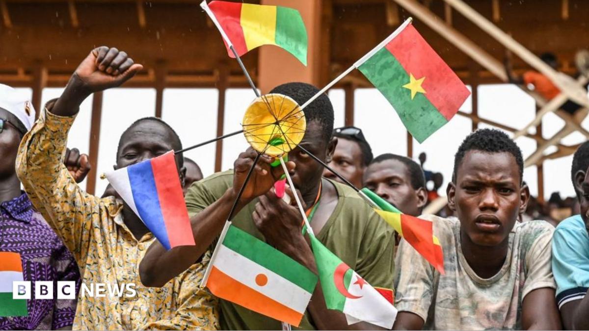 Man holding a round device that has the flags of Mali, Burkina Faso, Algeria, Niger and Russia attached to it. He is standing in a crowd and there are other people next to him.
