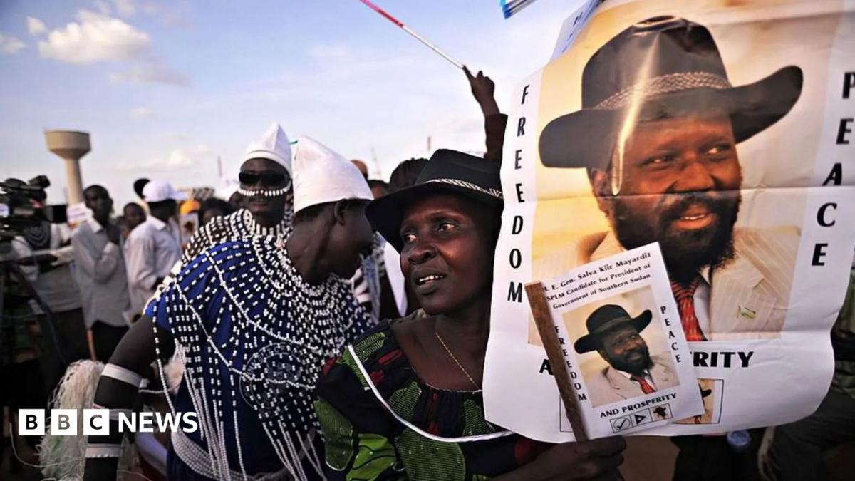 A crowd at a political rally holding a banner for South Sudan President Salva Kiir that reads "Freedom, peace and prosperity next to his portrait wearing a black cowboy hat