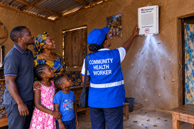 Community health worker in a blue vest demonstrates a medical device mounted on a mud-walled wall to a family of four in a rural home.