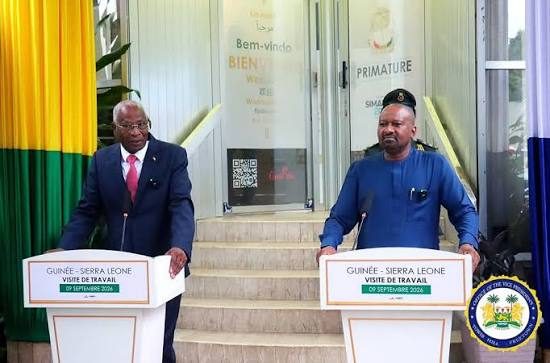 Two officials stand at separate podiums during a Guinea–Sierra Leone work visit, in front of a government building with welcome banners.