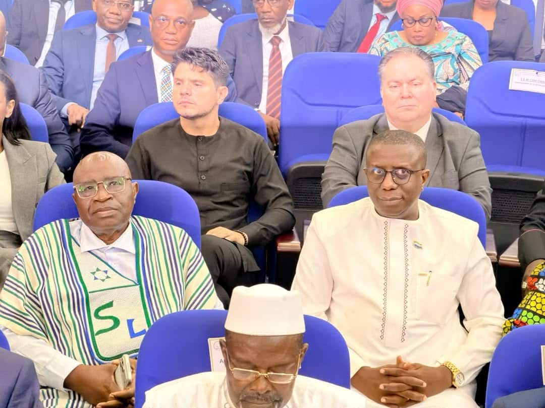 Front row of a formal event: a man wearing a white cap and glasses sits in the foreground next to a man in a white embroidered shirt, with other attendees in blue seats behind them.