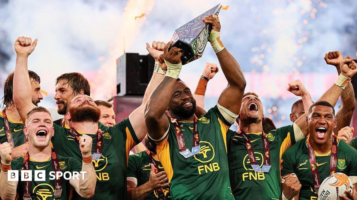 Siya Kolisi lifts the Rugby's Greatest Rivalry trophy, surrounded by happy team-mates in South Africa's largely green kit, as pyrotechnics light up the background
