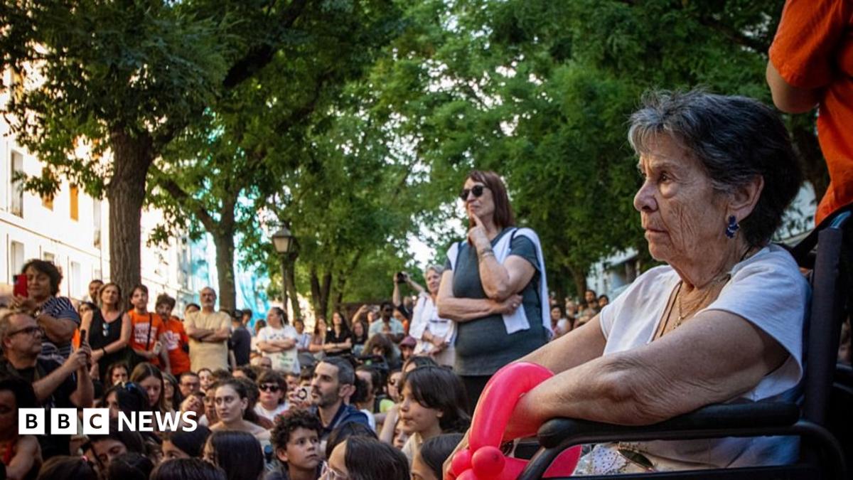 Maricarmen sits outdoors under some trees in a wheelchair. Behind her are a crowd of protesters