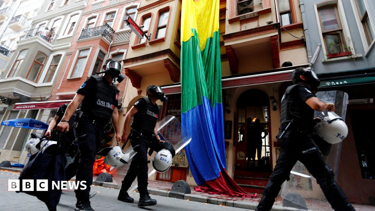 Riot police, with a rainbow flag in the background, chase LGBT activists as they try to gather for a pride parade, which was banned by the governorship in Istanbul. Officers are wearing black uniforms, wearing gas masks, and are carrying helmets and riot shields as they move through what appears to be a residential street.