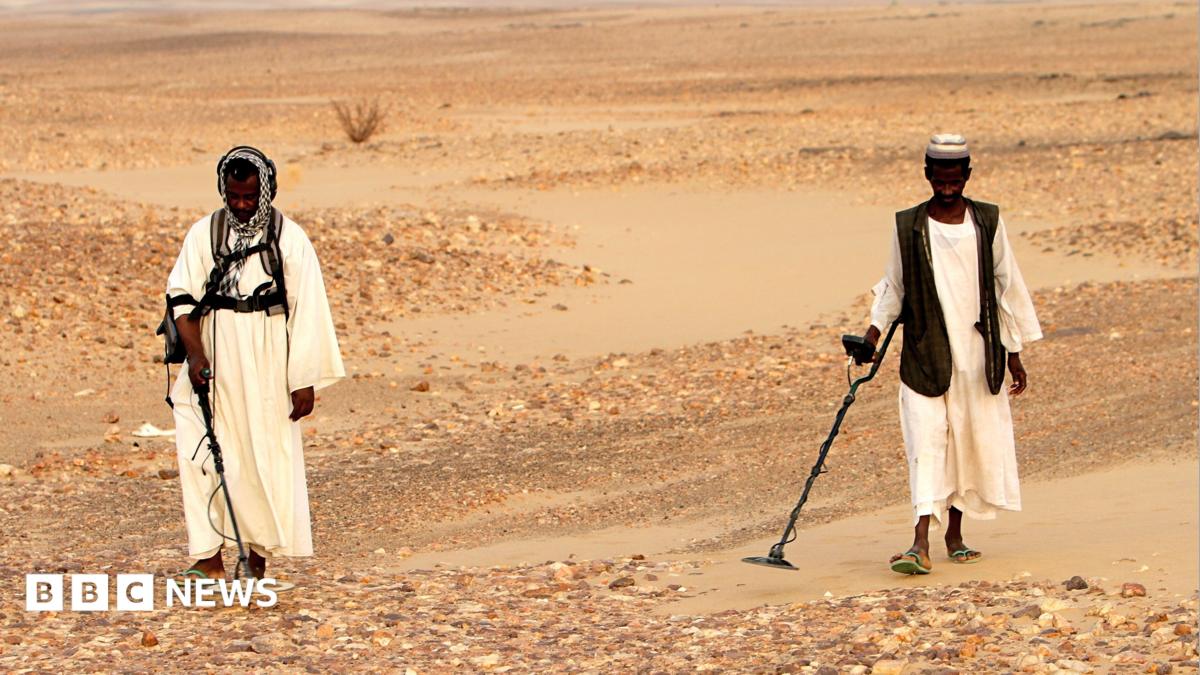 Sudanese men wearing traditional clothing search for gold with metal detectors in the desert.