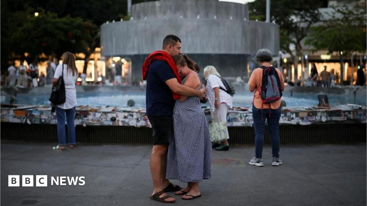 A man and woman embrace in front of the fountain at Dizengoff Square. The woman has her hand to her eyes as if she is crying.