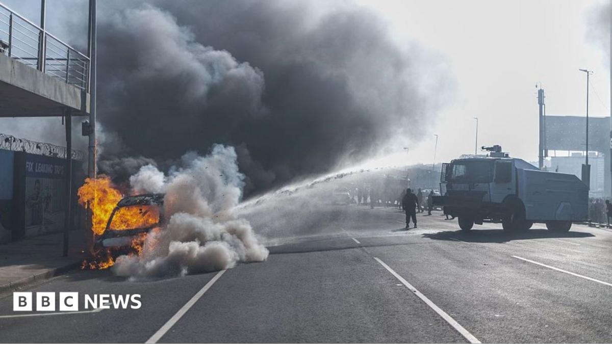 A vehicle burns on the side of a road as thick black smoke fills the air, while a police water-cannon truck sprays water towards the scene